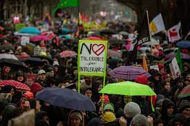 جمعیت زیادی از مردم با چتر در کنار هم ایستاده و تابلوهایی در دست دارند. یکی از آنها می گوید: "عدم تحمل برای عدم تحمل."